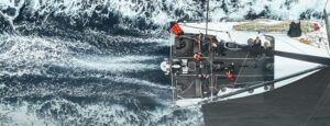 Top-down aerial view of a sailboat slicing through dark, foamy ocean with crew on deck