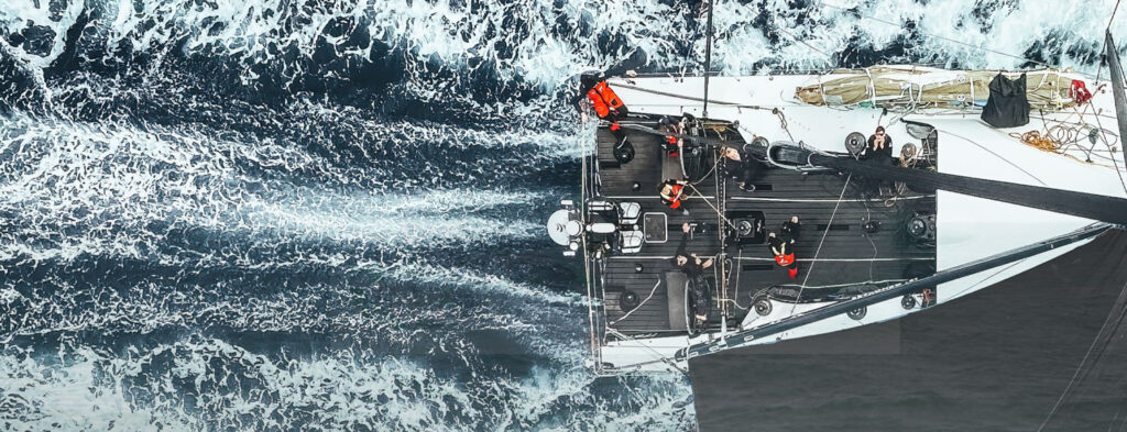 Aerial view of a sailboat cutting through blue water, white wake trailing behind as the crew works on deck.