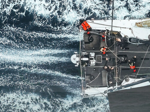 Aerial view of a sailboat cutting through blue water, white wake trailing behind as the crew works on deck.