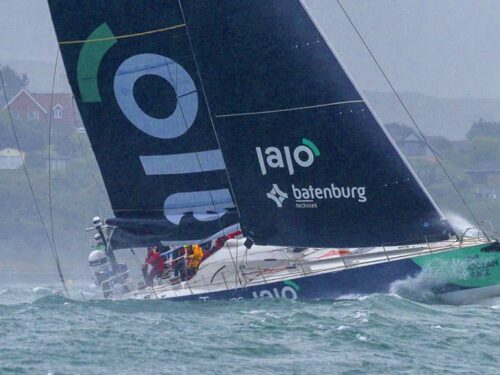 Race sailboat heeling sharply on rough water near a residential coastline, with crew on deck wearing life jackets and a dark sail bearing sponsor logos.