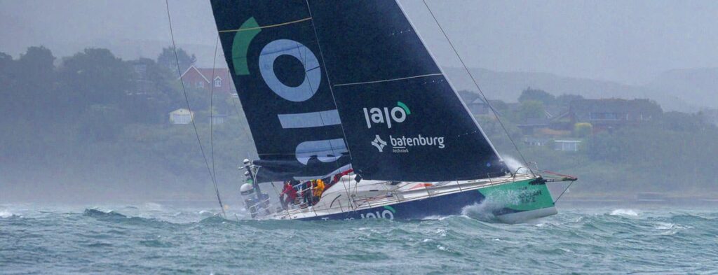 Sailing yacht tilts in choppy water near a coastline during a race, crew on deck adjusting lines as waves crash against hulls.